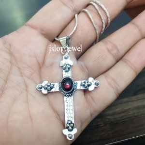 Puede incluir: Un colgante de cruz de plata con una piedra preciosa roja en el centro. La cruz tiene un diseño detallado con pequeñas cuentas alrededor de los bordes. El colgante está en una cadena de plata.