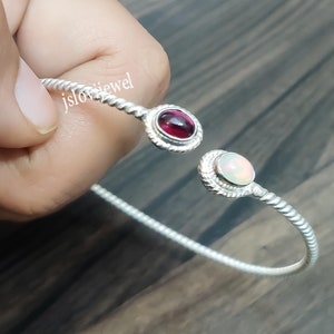 Puede incluir: Un brazalete de plata con un diseño trenzado. El brazalete tiene dos piedras redondas, una roja y una blanca.