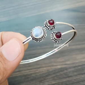 May include: A silver cuff bracelet featuring a large, round, iridescent moonstone and two smaller, round, deep red garnet gemstones. The stones are set in ornate, twisted silver metalwork. The text jslovijewel is visible on the bracelet.