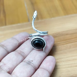 May include: A silver-toned ring in the shape of a snake, with a black oval stone at the base. The snake's head curves upwards. The ring is on a hand, with the brand name "jslovijewel" visible.