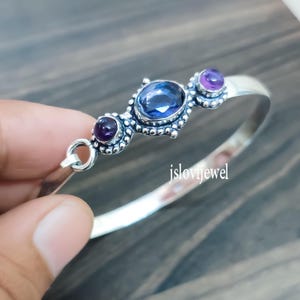 May include: A silver bangle bracelet with three gemstone accents. The center stone is a large oval blue gem, flanked by two smaller purple gems. The bracelet has a decorative beaded design around the gems. The text "jslovijewel" is visible.