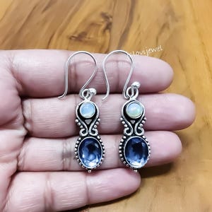 Op de afbeelding: Een paar zilveren oorhangers met blauwe en witte edelstenen. De oorbellen hebben een haaksluiting en hebben een ovale blauwe edelsteen aan de onderkant en een ronde witte edelsteen aan de bovenkant. Het ontwerp bevat decoratieve krullen.