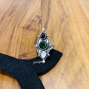 May include: A silver ring with an ornate design featuring three gemstones: black, green, and white. The ring is set against a wooden surface and a black fabric. The ring has the text "jslovijew" on the black fabric.