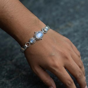 Könnte beinhalten: Ein silbernes Armband mit drei ovalen Mondsteinen, die in dekorativen Metallfassungen sitzen. Der mittlere Mondstein ist am größten und wird von zwei kleineren Steinen flankiert. Das Metallband hat eine polierte Oberfläche und eine glatte Textur.