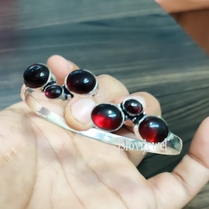 May include: Silver bangle bracelet with five red gemstone accents. The bracelet is open and has a smooth, polished finish.