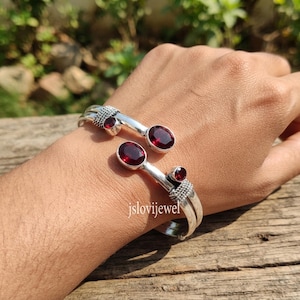 May include: A silver cuff bracelet featuring a double-band design with textured metal accents and four deep red oval-shaped gemstones. The bracelet has an open-ended style with two larger stones at the terminals and two smaller stones set along the bands.