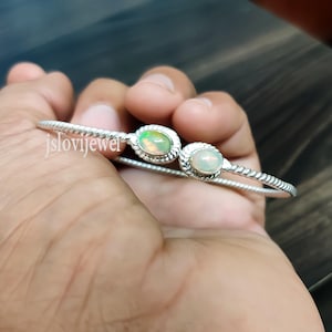 以下が含まれることがあります： 2つの楕円形のオパールストーンが付いたシルバーのバングルブレスレット。ブレスレットはねじれており、繊細なデザインです。