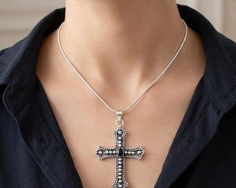 Puede incluir: Un collar con colgante de cruz en tono plateado que presenta una piedra ovalada negra central y detalles de cuentas intrincados en los bordes. La cruz cuelga de una cadena fina y lisa de tono plateado sobre una camisa de cuello oscuro.