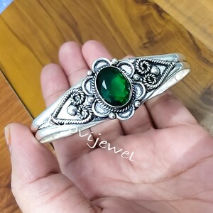 Op de afbeelding: Een zilveren manchetarmband met een ovale, smaragdgroene edelsteen. De armband heeft ingewikkelde filigraan- en kralen details rond de edelsteen en langs de band. Het woord "ovijewel" is zichtbaar.