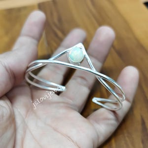 May include: A silver cuff bracelet with an opal gemstone. The bracelet has a layered design with a central V-shape and a round, iridescent opal. The bracelet is held in a hand.