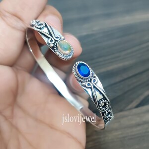 以下が含まれることがあります： 銀製のバングルブレスレット。オパールと青い宝石が、装飾的な銀のセッティングにセットされています。ブレスレットはオープンタイプで、繊細なデザインです。