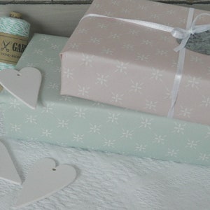 Puede incluir: Dos regalos envueltos en tonos pastel, rosa y verde menta, con un estampado floral blanco. El regalo rosa est&aacute; atado con una cinta blanca y una etiqueta de piedra gris. Etiquetas blancas en forma de coraz&oacute;n y un carrete de hilo completan la presentaci&oacute;n.