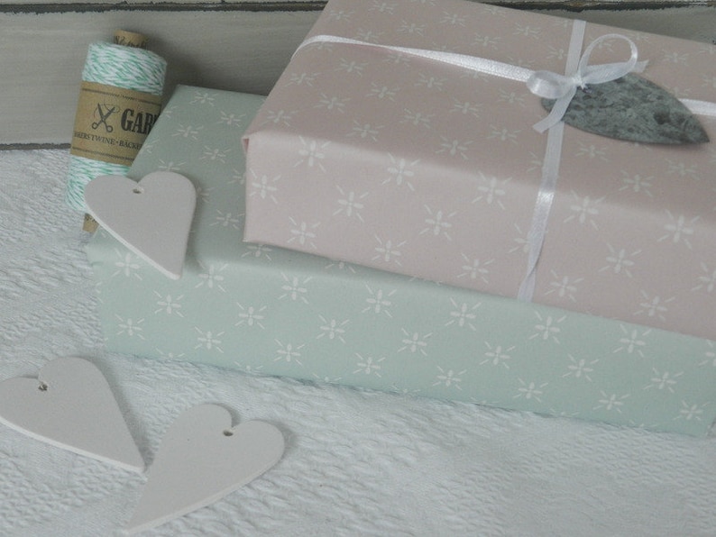 Puede incluir: Dos regalos envueltos en tonos pastel, rosa y verde menta, con un estampado floral blanco. El regalo rosa est&aacute; atado con una cinta blanca y una etiqueta de piedra gris. Etiquetas blancas en forma de coraz&oacute;n y un carrete de hilo completan la presentaci&oacute;n.