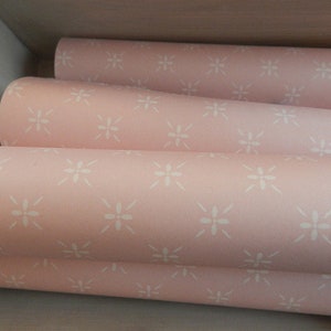 Puede incluir: Primer plano de varios rollos de papel pintado rosa con un dise&ntilde;o floral blanco repetido. Los rollos est&aacute;n apilados, revelando el patr&oacute;n y la textura. El papel pintado tiene una suave combinaci&oacute;n de colores pastel, ideal para la decoraci&oacute;n de interiores.