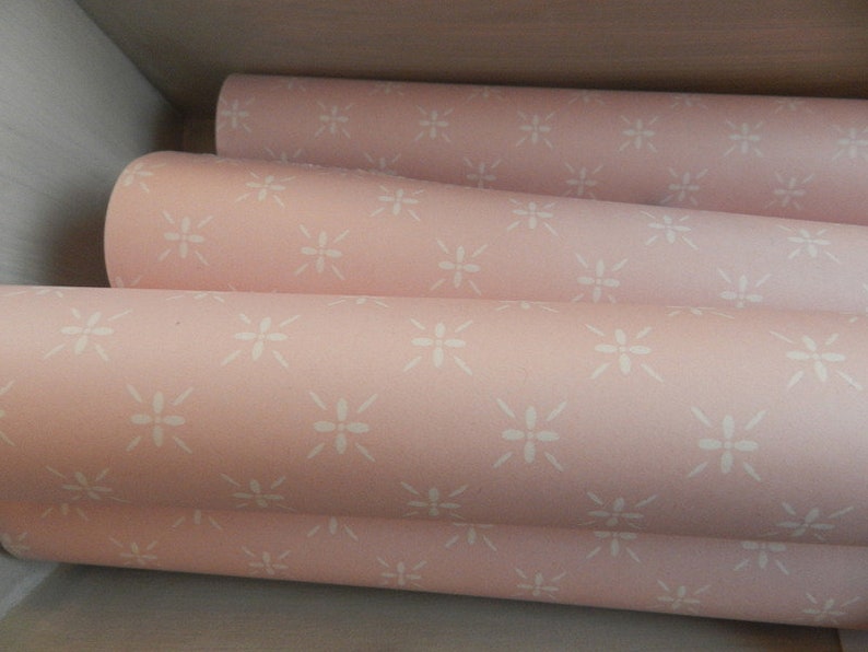 Puede incluir: Primer plano de varios rollos de papel pintado rosa con un dise&ntilde;o floral blanco repetido. Los rollos est&aacute;n apilados, revelando el patr&oacute;n y la textura. El papel pintado tiene una suave combinaci&oacute;n de colores pastel, ideal para la decoraci&oacute;n de interiores.