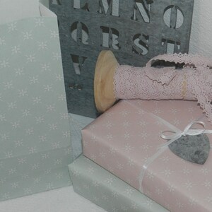 Puede incluir: Una colecci&oacute;n de materiales para envolver regalos. Incluye bolsas de papel azul claro y regalos envueltos en papel rosa y verde claro, todos con un estampado floral blanco. Una bobina de madera contiene cinta de encaje rosa. Un letrero de metal gris con letras est&aacute; en el fondo.