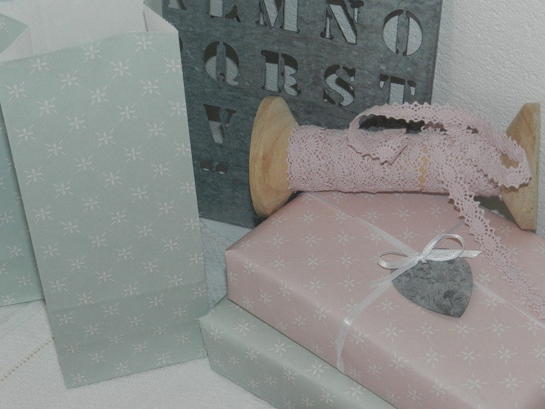 Puede incluir: Una colecci&oacute;n de materiales para envolver regalos. Incluye bolsas de papel azul claro y regalos envueltos en papel rosa y verde claro, todos con un estampado floral blanco. Una bobina de madera contiene cinta de encaje rosa. Un letrero de metal gris con letras est&aacute; en el fondo.