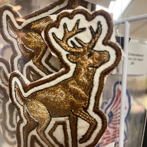 May include: Embroidered patch featuring a brown deer with antlers, outlined in white and brown. The patch is on a white background. The patch is an iron-on patch.