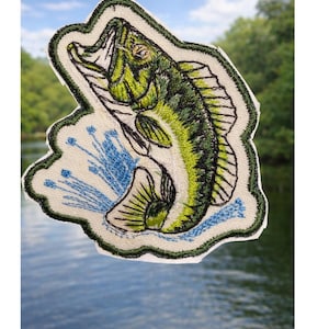 May include: Embroidered patch featuring a leaping largemouth bass. The fish is detailed with green and black shading, with blue water splashes. The patch has a white border and is set against a blurred background of water and trees.