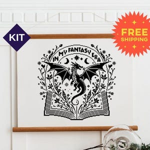 May include: A cross stitch pattern on white fabric featuring a black graphic of an open book with a dragon rising from the pages, surrounded by floral vines, stars, and the text "IN MY FANTASY ERA" arched across the top.