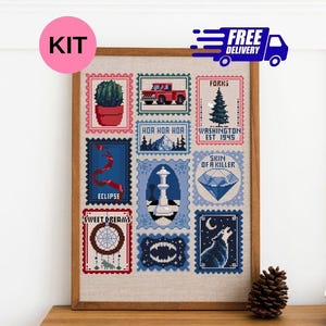 May include: A cross-stitch pattern framed in wood, featuring a grid of postage stamp-style illustrations including a red truck, a cactus, a pine tree, a chess piece, a diamond, a dreamcatcher, a wolf, and text like Forks and Eclipse.