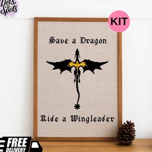 May include: A framed cross-stitch embroidery on light beige fabric featuring a black dragon silhouette with gold accents in the centre. The text "Save a Dragon" is stitched in black above the dragon, and "Ride a Wingleader" is stitched below.
