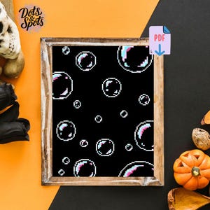 May include: A framed cross-stitch pattern featuring pixelated bubbles on a black background. The bubbles have white outlines and pink, blue, and white highlights. The frame is a light brown color. The image also includes the text "Dots & Spots" and a PDF icon.