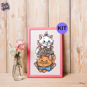 May include: A cross stitch kit featuring a vertical stack of three cartoon kittens in a pink frame. The top kitten is white with a pink bow, the middle is gray, and the bottom is orange with a blue bow. A purple circle with the text KIT is visible.