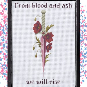 May include: A framed cross-stitch embroidery featuring a central dagger with a light-coloured hilt and a pink blade, surrounded by red poppy flowers and green leaves. The text "From blood and ash" and "we will rise" is stitched in black thread.