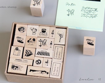 May include: A collection of wooden stamps with various cafe-themed illustrations, including coffee cups, pastries, and cafe tables. The stamps are arranged in a cardboard box, with a few individual stamps scattered around. The text "Wooden stamp" is visible.