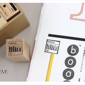 Puede incluir: Un sello de madera con las palabras "Reading List" y un gr&aacute;fico de libros. El sello est&aacute; junto a una p&aacute;gina de revista con la palabra "boo" y otro texto. Una caja de sellos est&aacute; en el fondo.