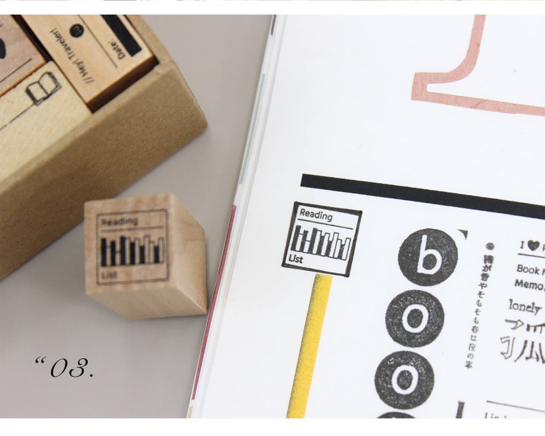 Puede incluir: Un sello de madera con las palabras "Reading List" y un gr&aacute;fico de libros. El sello est&aacute; junto a una p&aacute;gina de revista con la palabra "boo" y otro texto. Una caja de sellos est&aacute; en el fondo.