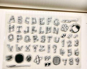 May include: Clear acrylic stamps featuring the alphabet, numbers, and various doodle designs. The letters and numbers are outlined in black, with a hand-drawn aesthetic. Includes punctuation marks and small graphic elements for creative projects.