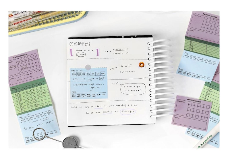 May include: A spiral-bound notebook with a black cover and white pages, surrounded by colorful sticky notes. The notebook has handwritten notes and the word "HAPPY!" on the top. The sticky notes are in shades of purple, green, and blue.
