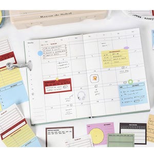 May include: An open planner with a calendar layout, surrounded by colorful sticky notes and a metal clip. The notes are in various colors, including red, yellow, and blue, with handwritten text. A pen case is visible in the background.