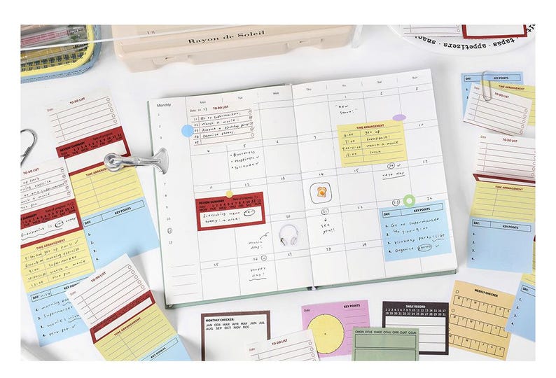May include: An open planner with a calendar layout, surrounded by colorful sticky notes and a metal clip. The notes are in various colors, including red, yellow, and blue, with handwritten text. A pen case is visible in the background.