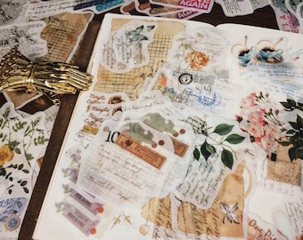 Pode incluir: Uma coleção de adesivos transparentes de estilo vintage com designs florais e botânicos, incluindo texto escrito à mão, e alguns com bolinhas. Os adesivos estão dispostos em um caderno com capa marrom.