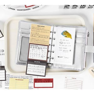 May include: A silver planner with a snap closure, open to reveal lined pages and calendar inserts. Surrounding the planner are various sticky notes, a small eraser, and a tray. The text "we rean what we sow" is visible on the eraser.