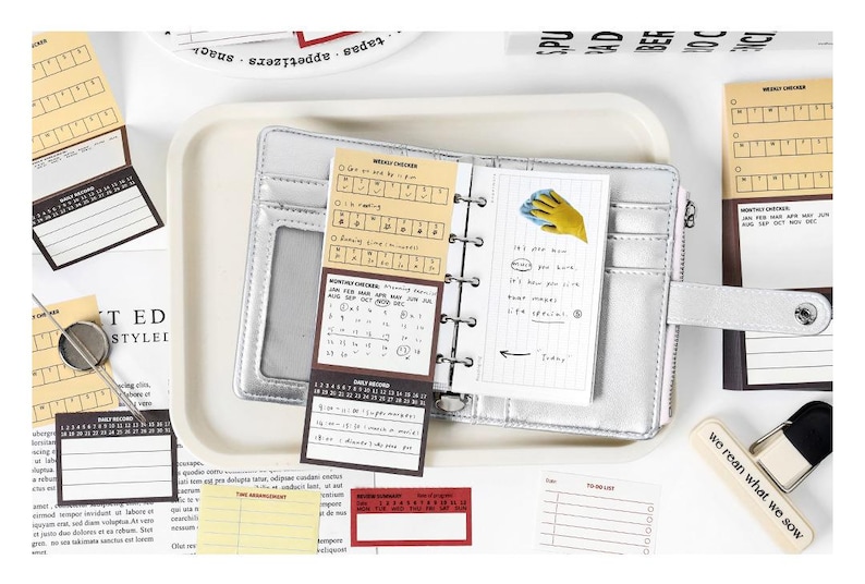 May include: A silver planner with a snap closure, open to reveal lined pages and calendar inserts. Surrounding the planner are various sticky notes, a small eraser, and a tray. The text "we rean what we sow" is visible on the eraser.