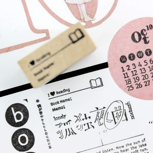 Puede incluir: Un collage de elementos de papeler&iacute;a con un sello de madera, un calendario rosa de octubre y texto japon&eacute;s. El sello tiene un icono de libro y texto. El tema es la lectura y el diario.