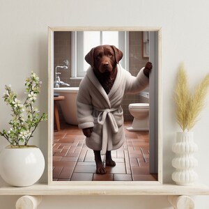 Könnte beinhalten: Gerahmter Druck mit einem braunen Labrador Retriever in einem weißen Bademantel, der sich in einem Badezimmer befindet. Der Hund lehnt an einem Türrahmen. Das Badezimmer hat eine Badewanne, eine Toilette und einen braunen Fliesenboden.
