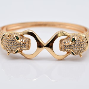 Könnte beinhalten: Goldfarbenes Armband mit zwei Pantherköpfen mit smaragdgrünen Augen und besetzt mit glitzernden Kristallen. Das Armband hat ein Scharnierdesign und ist wahrscheinlich aus goldfarbenem Metall gefertigt. Die Pantherköpfe sind einander zugewandt.