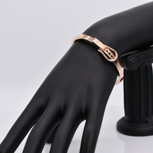 Op de afbeelding: Een ros&eacute;gouden armband met een gespsluiting. De armband is op een zwarte mannequinhand.