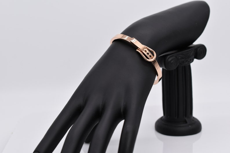 Op de afbeelding: Een ros&eacute;gouden armband met een gespsluiting. De armband is op een zwarte mannequinhand.