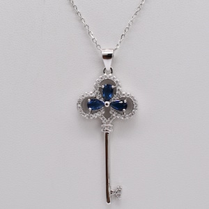 May include: A silver-toned pendant necklace featuring a key design. The key's head is a clover shape with three dark blue, pear-cut gemstones surrounded by small, clear stones. The key's stem and bow are also detailed with clear stones.