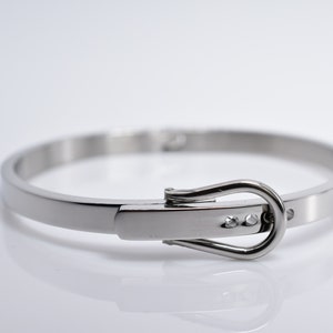 Op de afbeelding: Een zilveren armband met een gespsluiting. De gesp heeft drie kleine, heldere stenen.