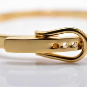 Op de afbeelding: Een gouden armband met een gespsluiting. De gesp is simpel, rond en heeft drie kleine gaatjes.