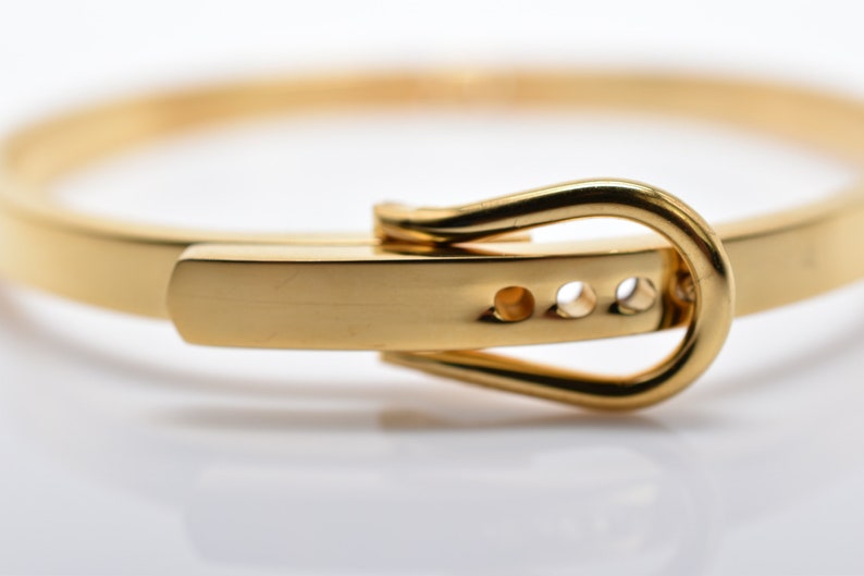 Op de afbeelding: Een gouden armband met een gespsluiting. De gesp is simpel, rond en heeft drie kleine gaatjes.