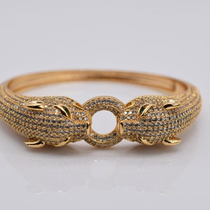 Könnte beinhalten: Goldfarbenes Armreif-Armband mit zwei Pantherköpfen, die mit kleinen, funkelnden Steinen besetzt sind. Das Armband hat ein Scharnierdesign und einen kreisförmigen Verschluss. Die Pantherköpfe haben grüne Augen.