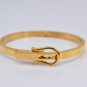 Op de afbeelding: Een gouden armband met een gespsluiting. De gesp heeft drie kleine, heldere stenen.